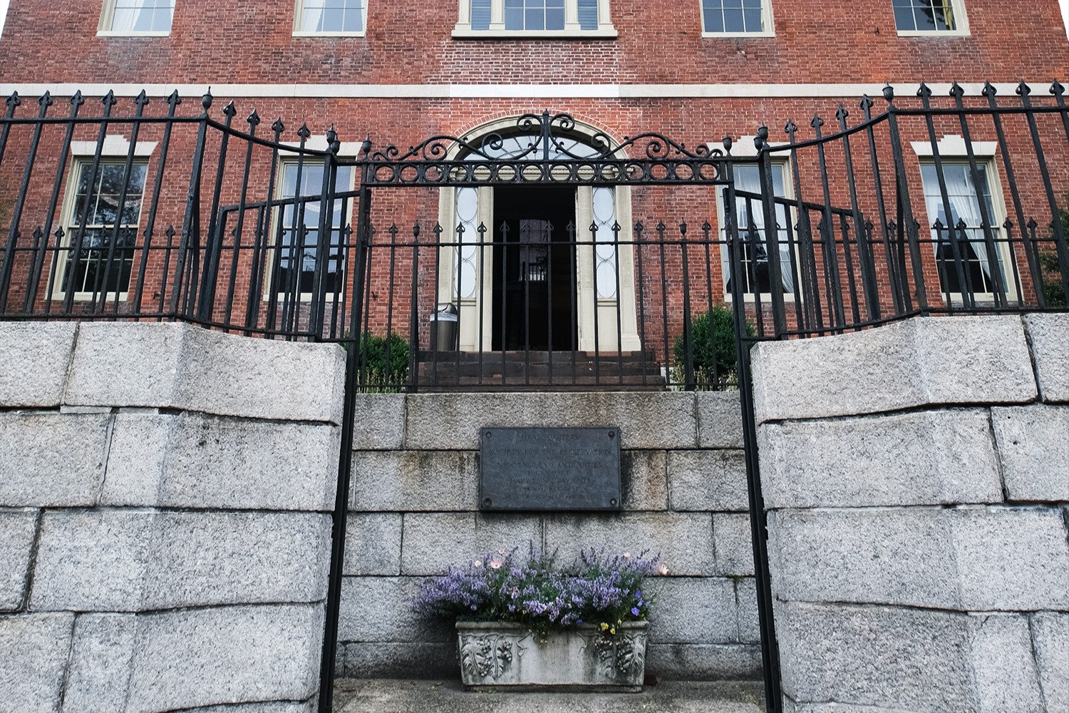 Exterior facade of a red brick mansion behind a cast iron gate around grey stone terrace.